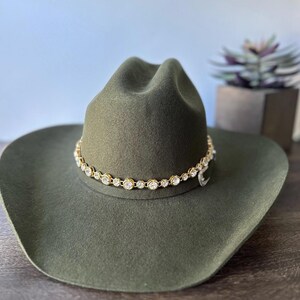 Pu&ograve; includere: Un cappello da cowboy in feltro verde oliva caratterizzato da una fascia a catena color oro decorata con cristalli trasparenti, rotondi e sfaccettati che circondano la corona. Il cappello presenta una corona strutturata e una tesa larga.