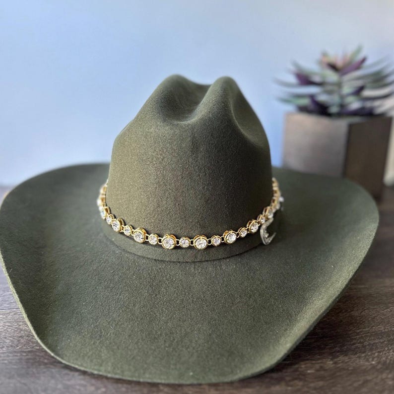 Pu&ograve; includere: Un cappello da cowboy in feltro verde oliva caratterizzato da una fascia a catena color oro decorata con cristalli trasparenti, rotondi e sfaccettati che circondano la corona. Il cappello presenta una corona strutturata e una tesa larga.