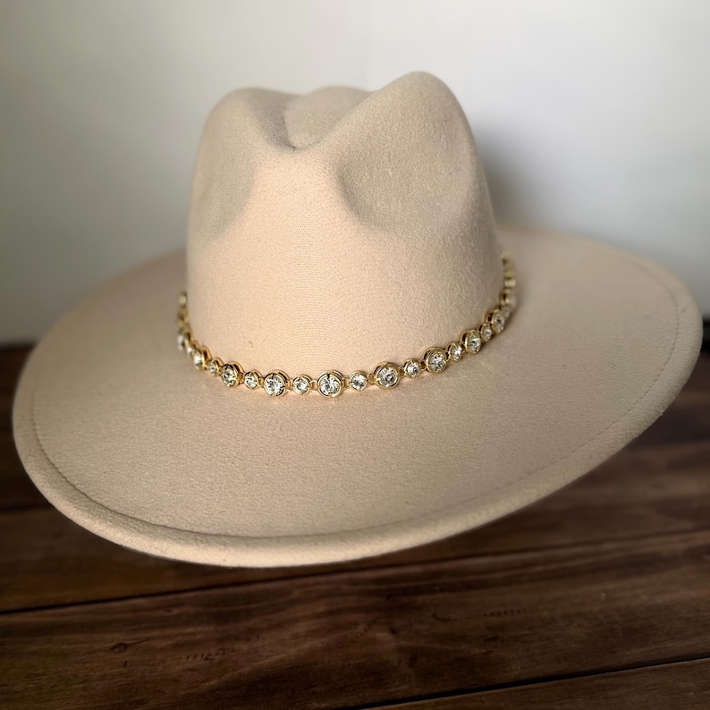 Pu&ograve; includere: Un cappello fedora beige con tesa larga e corona pizzicata. La base della corona &egrave; circondata da una fascia color oro decorata con una fila continua di cristalli trasparenti, rotondi e sfaccettati, incastonati in castoni metallici.