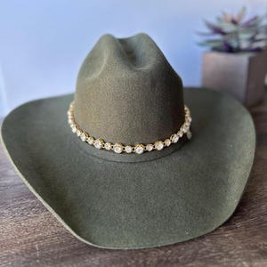Pu&ograve; includere: Un cappello da cowboy in feltro verde oliva caratterizzato da una fascia a catena color oro decorata con una serie di cristalli trasparenti, rotondi e sfaccettati che circondano la corona. Il cappello ha una tesa larga e curva e una piega centrale.