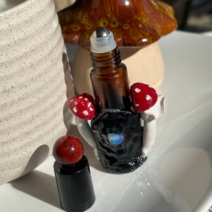 May include: Two small glass bottles with roller balls. The bottles are filled with a dark liquid. The bottles are decorated with red and white mushroom-shaped charms. The bottles are sitting on a white surface.