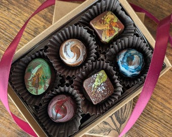 May include: A rectangular cardboard box containing six assorted artisan chocolates in individual dark brown paper liners. The chocolates feature various shapes and colorful, marbled, or speckled finishes in shades of green, brown, blue, and red, tied with a burgundy ribbon.