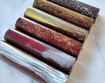 May include: Six chocolate bars with different colored coatings. The bars are arranged in a row from left to right, with the darkest bar on the left and the lightest bar on the right. The bars are all about the same size and shape, and they are all wrapped in a clear plastic.