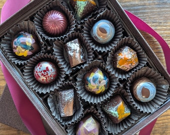 May include: A square box containing twelve assorted artisan chocolates in individual paper liners. Each chocolate features a unique glossy finish with varied colours including metallic gold, blue, red, purple, and brown, showcasing intricate patterns.