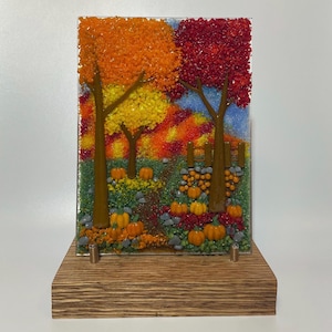 May include: A rectangular fused glass art piece depicting an autumn forest scene with orange, yellow, and red trees, a dirt path, and orange pumpkins scattered on the ground. The glass panel is supported by a rectangular wooden base stand.