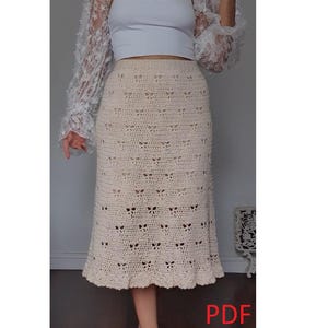 Puede incluir: Falda de crochet color crema, hasta la rodilla, con dobladillo festoneado. La falda presenta un patrón calado repetitivo. También se ven una camiseta sin mangas blanca y una chaqueta de encaje blanco con mangas con volantes.