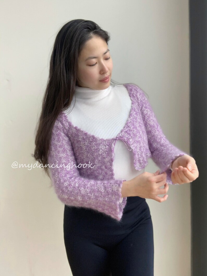 May include: A purple and white fuzzy cropped cardigan with a white turtleneck underneath. The cardigan has a V-neck and a tie at the front. The text "@mydancinghook" is visible in the bottom left corner of the image.