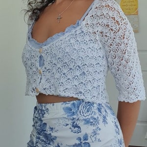 May include: A white crocheted crop top with a button closure and a floral print skirt. The top has a scalloped edge and the skirt has a blue and white floral pattern.