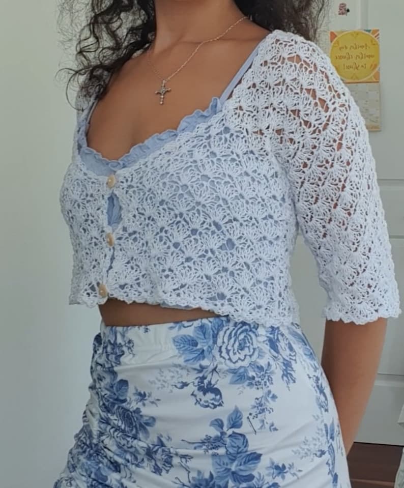 May include: A white crocheted crop top with a button closure and a floral print skirt. The top has a scalloped edge and the skirt has a blue and white floral pattern.