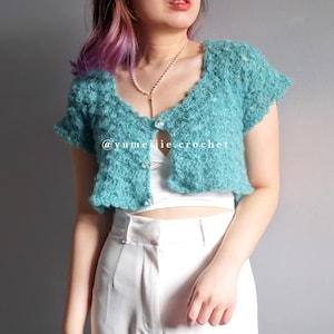 May include: A teal blue crocheted cropped cardigan with a button closure. The cardigan has a scalloped edge and short sleeves. The model is wearing the cardigan over a white tank top and white pants. The text "@yumeijie.crochet" is visible on the cardigan.