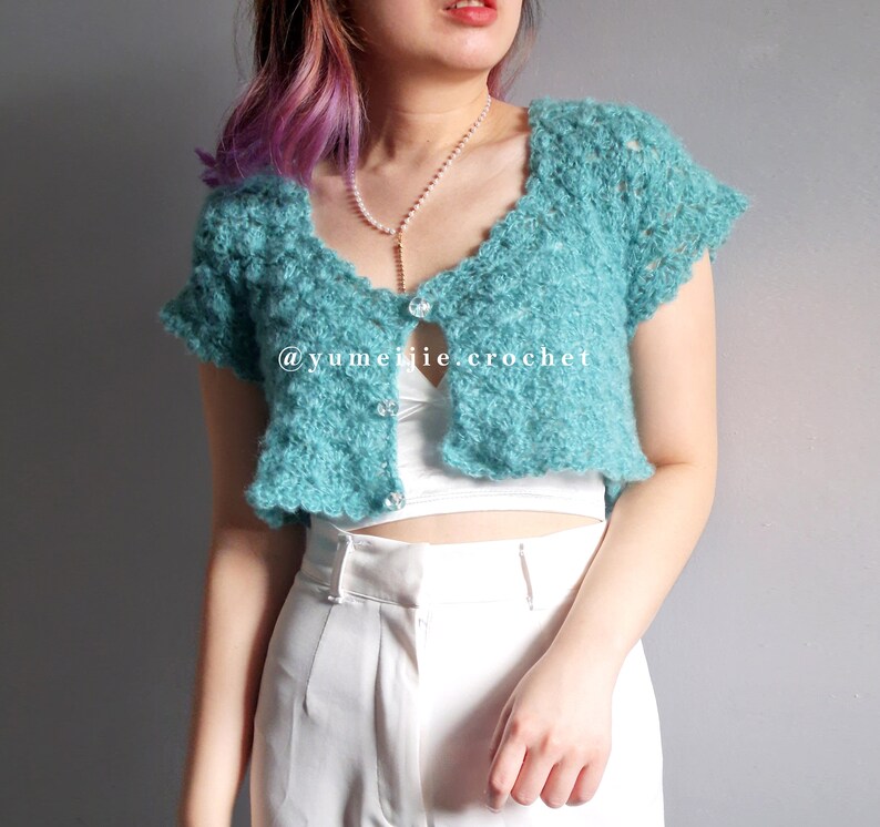 May include: A teal blue crocheted cropped cardigan with a button closure. The cardigan has a scalloped edge and short sleeves. The model is wearing the cardigan over a white tank top and white pants. The text "@yumeijie.crochet" is visible on the cardigan.