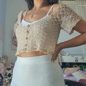 May include: A beige crochet crop top with a button closure and short sleeves. The top has a delicate, lacy pattern and a scalloped hem.
