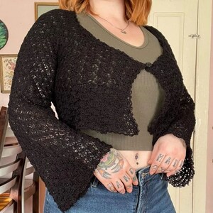 May include: Black crochet cardigan with a loose fit and bell sleeves. The cardigan is open in the front and has a single button closure.