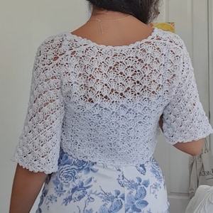 May include: White crocheted cropped cardigan with a delicate openwork pattern. The cardigan has a scoop neckline and short sleeves.