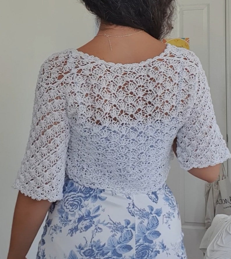 May include: White crocheted cropped cardigan with a delicate openwork pattern. The cardigan has a scoop neckline and short sleeves.