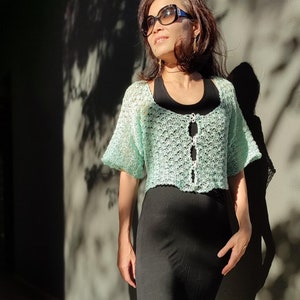 May include: A woman wearing a light green crocheted crop top with a black skirt and sunglasses.