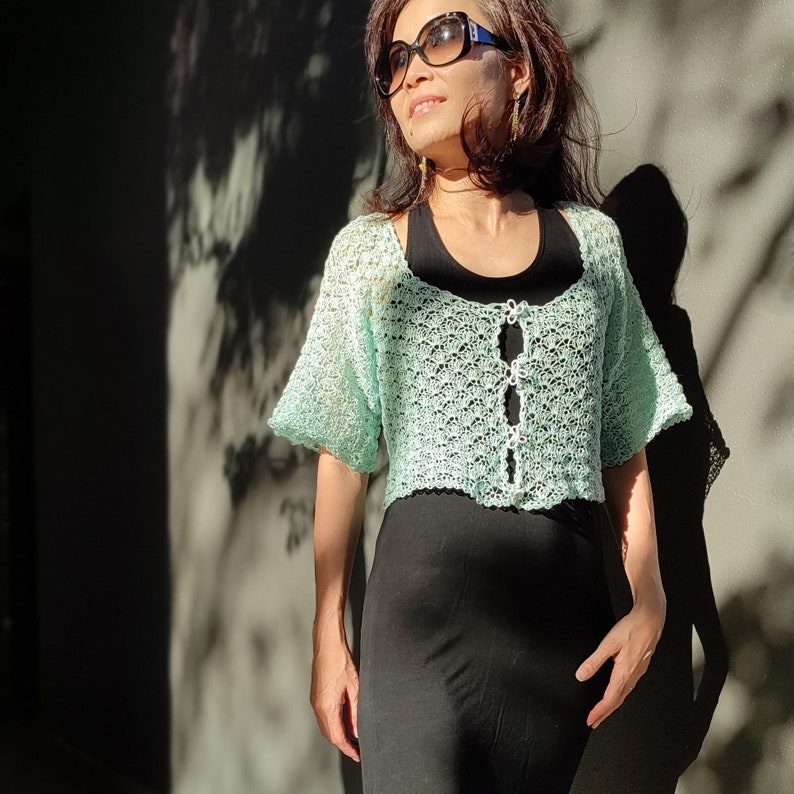May include: A woman wearing a light green crocheted crop top with a black skirt and sunglasses.