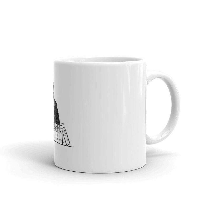 May include: A white coffee mug with a black and white illustration of a person sitting at a desk with a stack of books. The person is wearing a suit and tie. The text on the books reads 'MILL ST.'