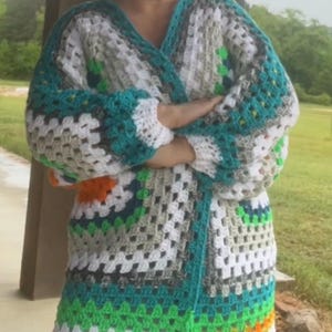 May include: A handmade, open-front crochet cardigan in a granny square pattern. The color scheme includes white, teal, gray, green, and orange. The long-sleeved garment is knee-length and has a V-neck.