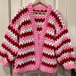 May include: A handmade, open-front crochet cardigan in a vibrant color scheme. The sweater features alternating stripes of red, white, and pink, with a solid pink border and collar. The sleeves are long and voluminous.