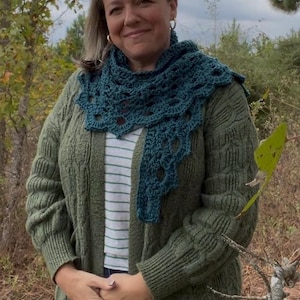 May include: A woman wearing a teal crocheted shawl over a green cardigan. The shawl has a scalloped edge and openwork design. The cardigan is a cable knit style. The woman is also wearing a striped shirt.