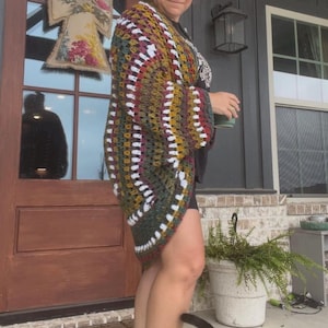 May include: A colorful, handmade crochet cardigan in shades of green, yellow, red, and white. The open-front cardigan has a geometric pattern and is worn by a person standing on a porch. A cat is in the foreground.