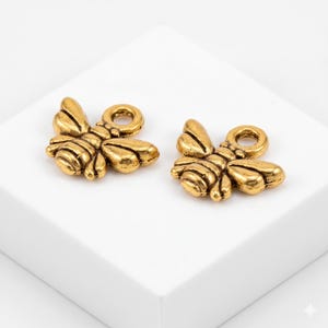 May include: Two gold-colored bee charms with detailed wings and body segments. Each charm has a small loop at the top for attaching to jewelry or other crafts. The bees are positioned on a white surface.