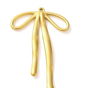 Può includere: Un ciondolo in metallo color oro a forma di fiocco. Il ciondolo ha un anello nella parte superiore per appenderlo e due elementi lunghi, curvi, a forma di goccia che pendono sotto il fiocco. Il metallo ha una finitura opaca.