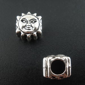 May include: A silver-toned metal bead shaped like a sun with a face. The bead features a raised, detailed sun design on the front and a circular hole running through the center for stringing. The metal has an antiqued finish with dark recessed areas.