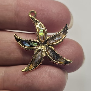 May include: A gold-colored starfish pendant with iridescent abalone shell inserts. The starfish has five points, each with a shell inlay. The pendant has a small loop at the top for attaching to a chain or cord. The edges are detailed.