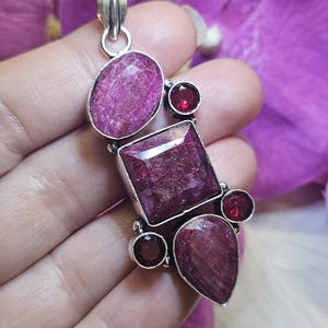 May include: A silver pendant featuring a cluster of faceted, deep red gemstones. The pendant includes an oval, square, and teardrop-shaped ruby, along with smaller round garnets. The gemstones are set in a silver metal frame.