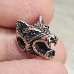 May include: A metallic silver-toned bead shaped like a snarling wolf head with detailed fur textures, pointed ears, and an open mouth showing teeth. The bead features a circular hole on the side for threading, with a polished finish and dark accents.