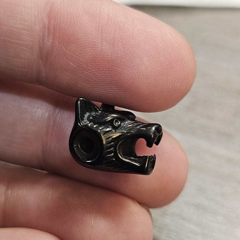 May include: A small, dark metallic bead shaped like a wolf head with a snarling expression, featuring detailed fur textures and an open mouth. The bead has a circular hole on the side, designed for threading onto a cord or chain for jewelry making.