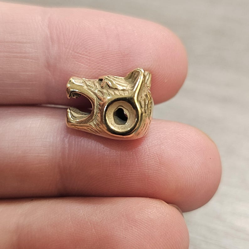 May include: A small, gold-colored metal bead shaped like the head of a roaring bear. The bead features detailed fur textures, a wide-open mouth, and a circular hole passing through the side for threading onto a cord, chain, or wire.
