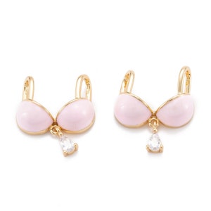 May include: A pair of gold-tone earrings with pink enamel and a clear crystal drop. The earrings are shaped like a bra.