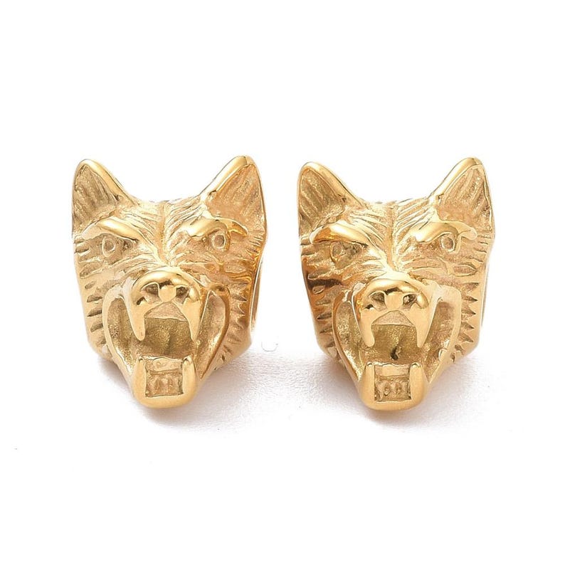 May include: A pair of gold-toned metal stud earrings shaped like the head of a snarling wolf. Each earring features detailed fur textures, pointed ears, and an open mouth revealing sharp teeth, with a polished finish on the metallic surface.