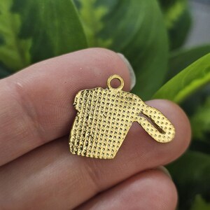 May include: A gold-coloured charm in the shape of a coffee cup with a handle. The charm has a textured surface with small, evenly spaced dots. A small loop is at the top for attaching to a chain or bracelet. The charm is held in a person's hand.