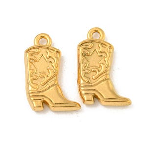 May include: Two gold-colored cowboy boot charms. Each charm features a star design and decorative scrollwork. The boots have a loop at the top for easy attachment to jewelry or other crafts. The charms are made of a shiny, metallic material.