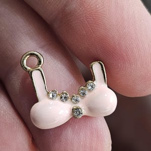 May include: A small, light pink and gold-colored charm in the shape of a bra. The charm is adorned with several clear, sparkling rhinestones. The charm has a gold loop at the top and is set against a blurred background.