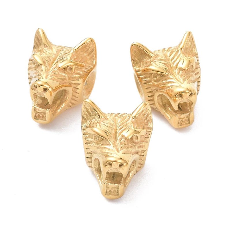 May include: Three gold-colored metal beads shaped like wolf heads with detailed fur textures and open mouths. Each bead features a polished finish and a hollow center, designed with a snarling expression and pointed ears, arranged in a triangular formation.