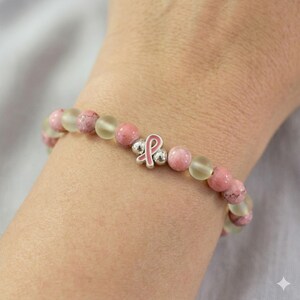 May include: A bracelet with pink and translucent beads, and a silver charm with a pink ribbon. The bracelet is displayed on a wrist, highlighting the colours and textures of the beads and charm. The beads are likely made of glass or stone.