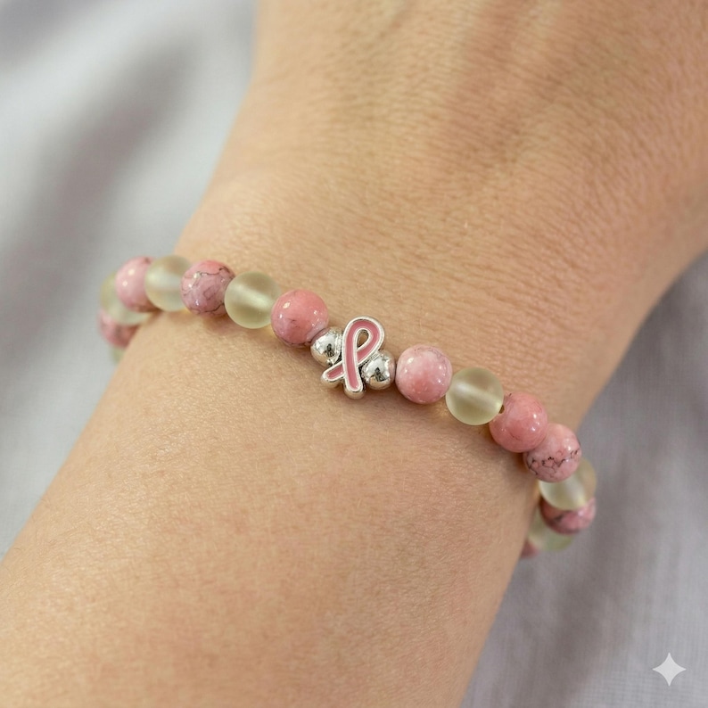 May include: A bracelet with pink and translucent beads, and a silver charm with a pink ribbon. The bracelet is displayed on a wrist, highlighting the colours and textures of the beads and charm. The beads are likely made of glass or stone.
