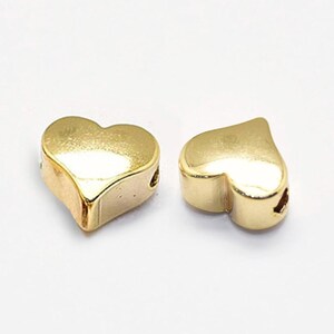 以下が含まれることがあります： 金色のハート型のビーズが2つ。各ビーズは滑らかで反射する表面と、紐を通すための穴があります。ビーズは白い背景に表示されています。これらのビーズは、ジュエリー作りに適しています。