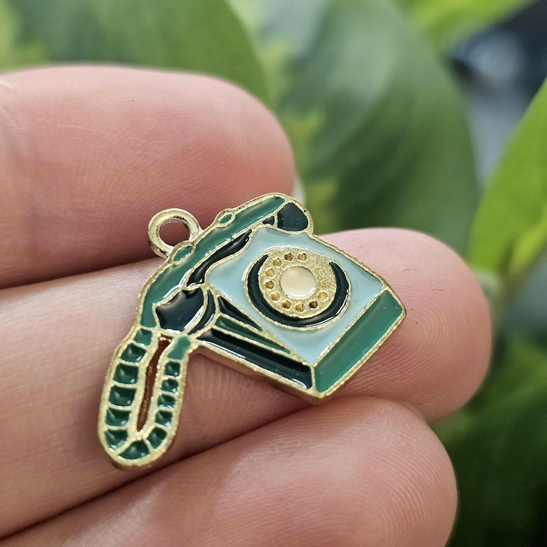 May include: A vintage telephone charm with a gold-coloured border. The charm features a light blue dial and receiver, with dark green and black accents. The rotary dial is cream-coloured. The charm is likely made of metal and enamel.