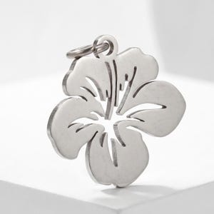 Puede incluir: Colgante de flor de hibisco plateado con un peque&ntilde;o lazo para una cadena. El colgante presenta un dise&ntilde;o recortado detallado, que muestra los p&eacute;talos y el centro de la flor. Hecho de un material met&aacute;lico brillante.