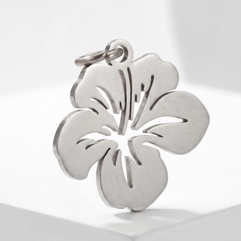 Puede incluir: Colgante de flor de hibisco plateado con un peque&ntilde;o lazo para una cadena. El colgante presenta un dise&ntilde;o recortado detallado, que muestra los p&eacute;talos y el centro de la flor. Hecho de un material met&aacute;lico brillante.
