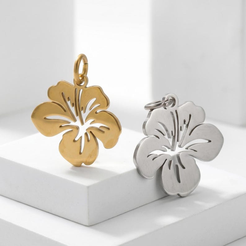 Puede incluir: Dos colgantes con forma de flor. Uno es dorado, el otro plateado. Ambos colgantes tienen un dise&ntilde;o de flor de hibisco con detalles recortados. Los colgantes se exhiben sobre una superficie blanca, mostrando sus intrincados patrones florales.