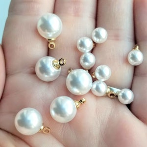 Puede incluir: Una colección de cuentas de perlas blancas con bucles de metal dorado. Las cuentas son de varios tamaños y están esparcidas en una mano.