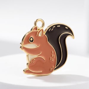 May include: A gold-colored metal squirrel charm with a brown body, black tail, and white accents. The charm has a loop at the top for attaching to a necklace or bracelet. The squirrel is in a sitting position.