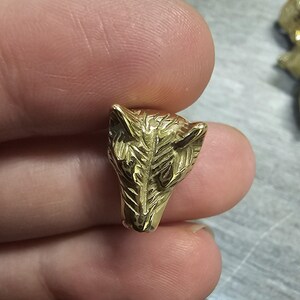 May include: A small, metallic gold-colored bead shaped like the head of a wolf. The surface features detailed, engraved lines that define the ears, snout, and facial structure of the animal, creating a textured and reflective finish on the metal.